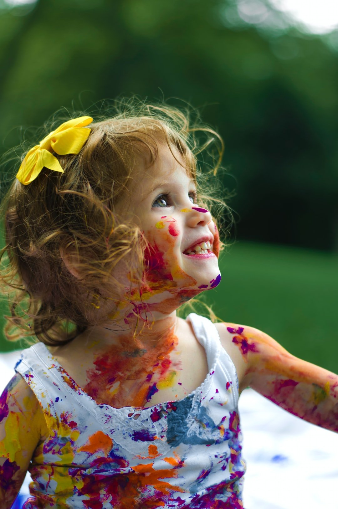 <p>Young girl smiling and splodged with different colours of paint.</p>